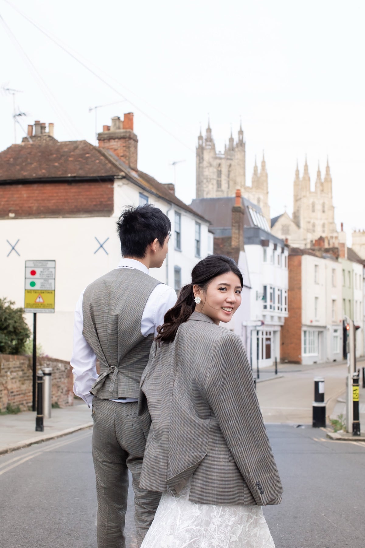 A couple wearing matching gray plaid wedding attire stands on a street in front of historic buildings and cathedral spires. The man is shown from the back wearing a gray plaid vest and trousers over a white shirt, while the woman looks back over her shoulder wearing a matching gray plaid blazer and a white lace dress.