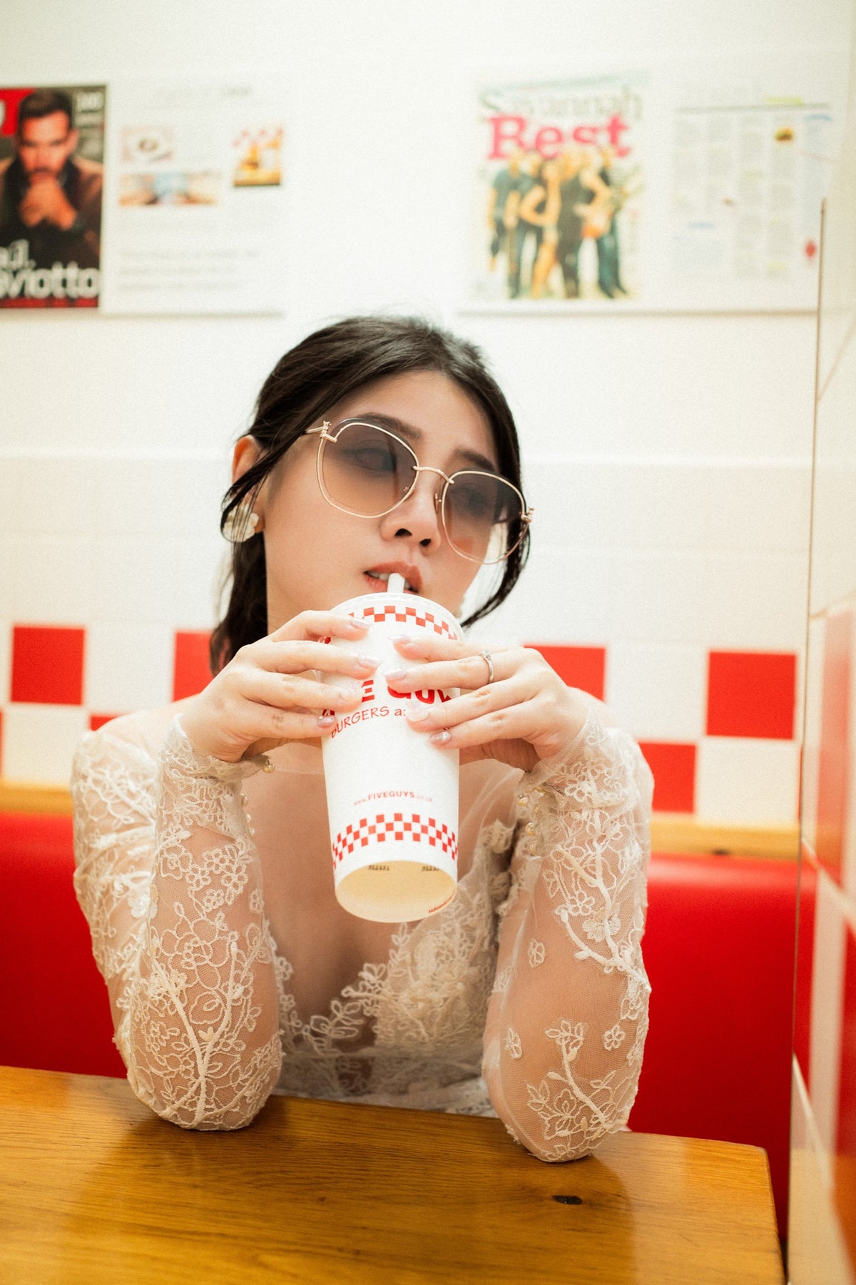 A woman wearing round sunglasses and a sheer white lace long-sleeve top sits in a red booth at a fast-food restaurant, holding a white paper drink cup with a red checkered pattern.