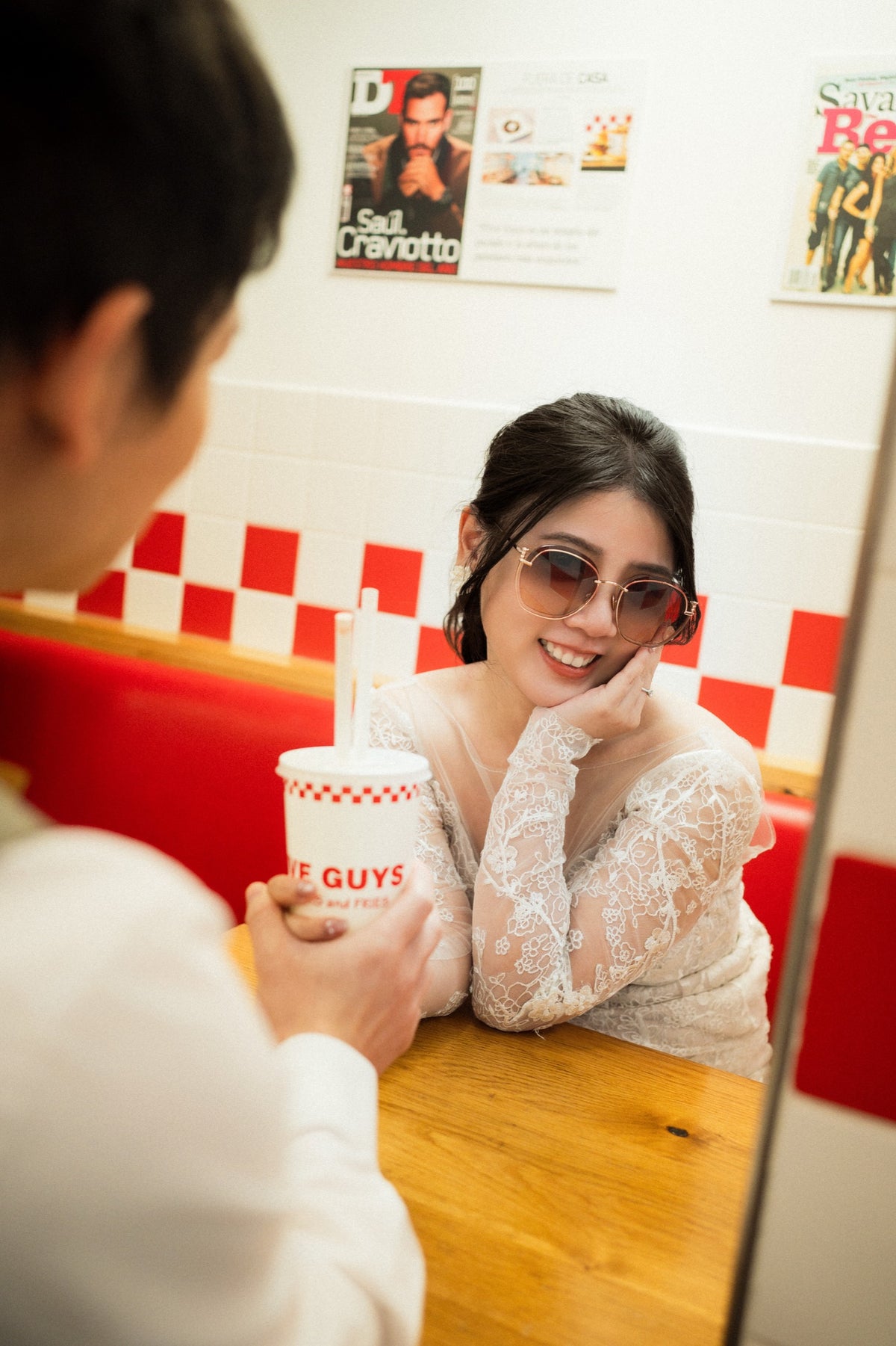 A woman in a white lace top and sunglasses smiles while sitting in a red and white checkered diner booth, holding a drink cup with the 'Invisible Girls' logo, with a man in the foreground.