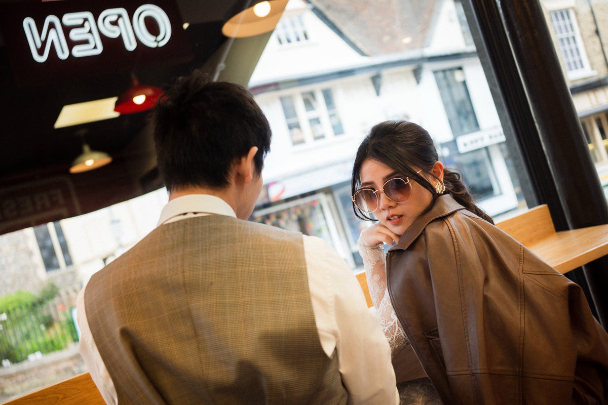 A woman wearing round sunglasses and a brown jacket sits at a cafe table talking to a man from behind, who is wearing a beige plaid vest.