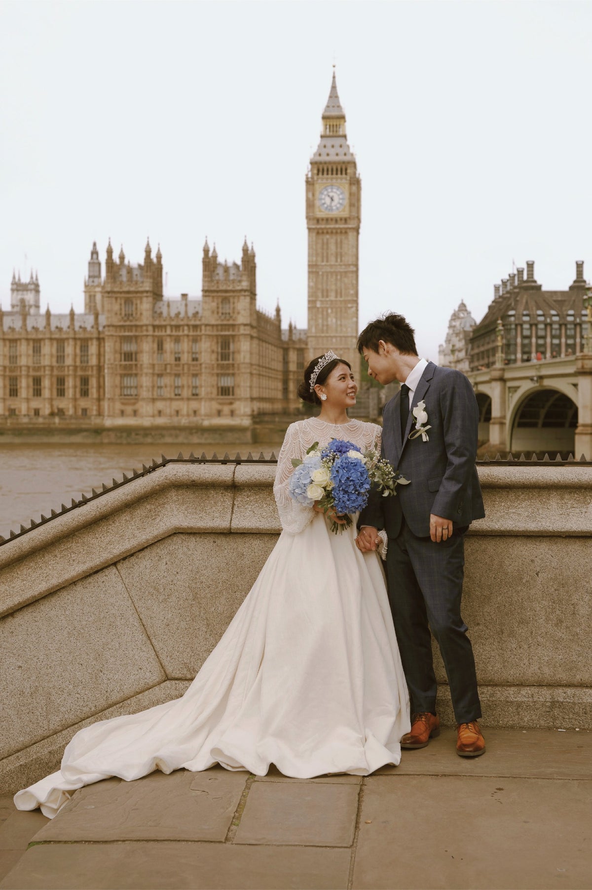 A bride in a white wedding gown and a groom in a dark suit pose together on a stone bridge in London, with Big Ben and the Houses of Parliament in the background.