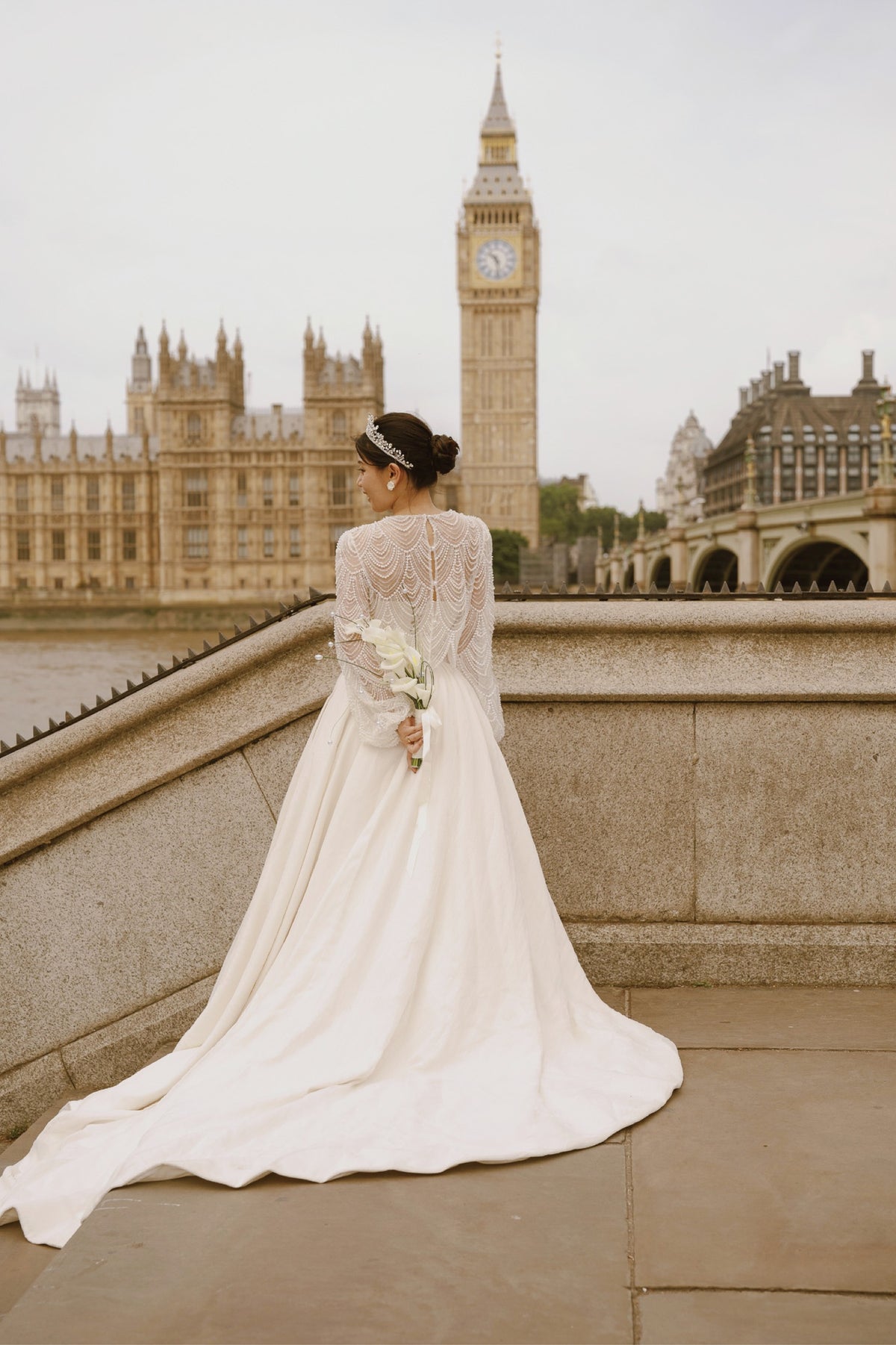 A bride in a long-sleeve lace wedding gown with a train stands on a stone bridge in London, facing Big Ben and the Houses of Parliament, holding a bouquet of white flowers.