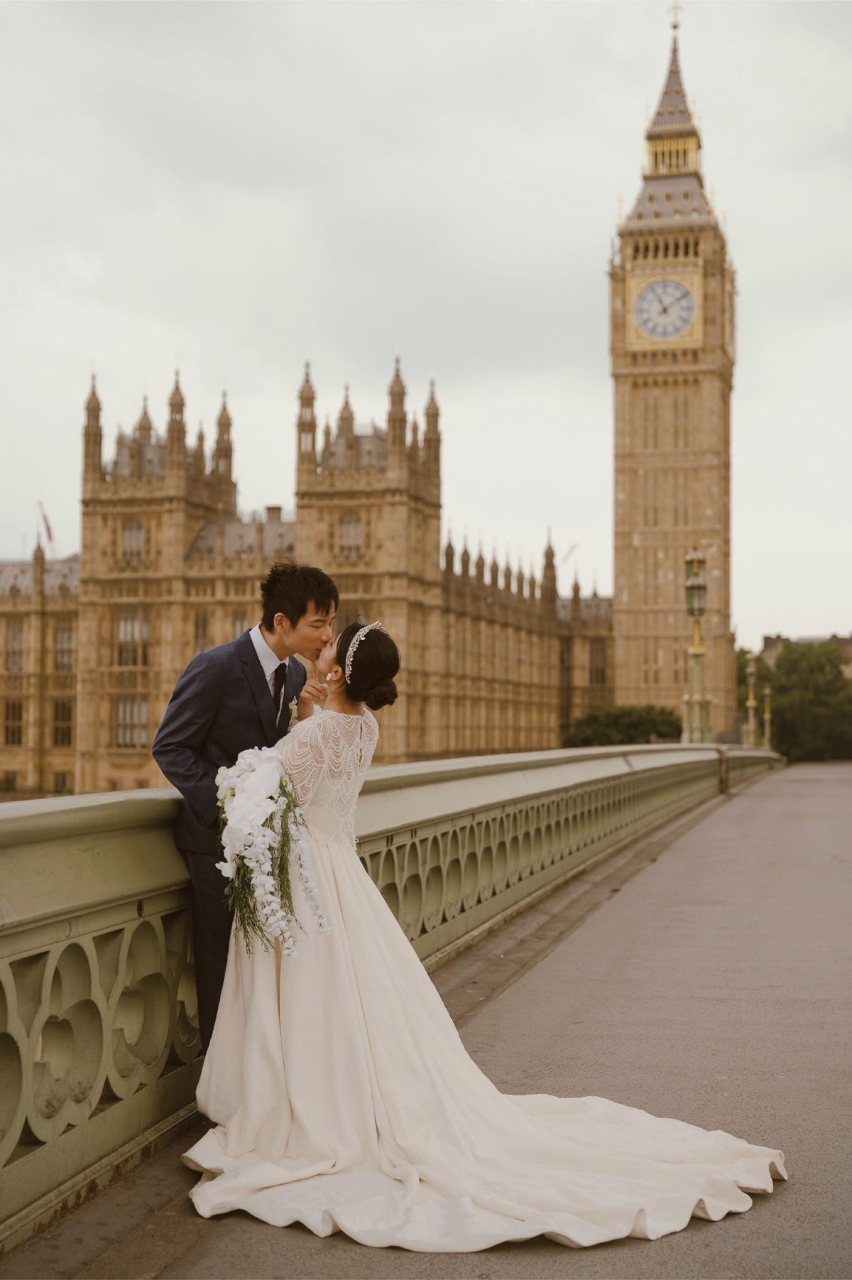 A bride in a long white wedding gown with a train and a groom in a dark suit kiss on a bridge, with Big Ben and the Houses of Parliament in London in the background.