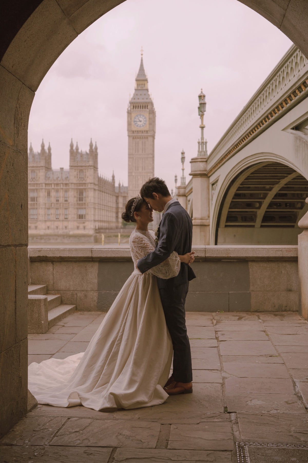 A bride in a long white wedding gown and a groom in a dark suit embrace and kiss under a stone archway, with Big Ben and the Houses of Parliament in London in the background.