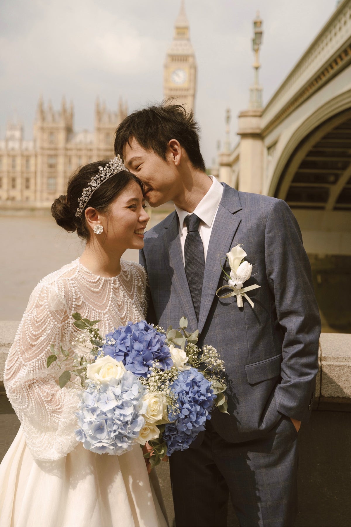 A bride in a white lace wedding dress and tiara holds a bouquet of blue and white flowers while standing next to a groom in a blue suit, with Big Ben and a bridge in the background.