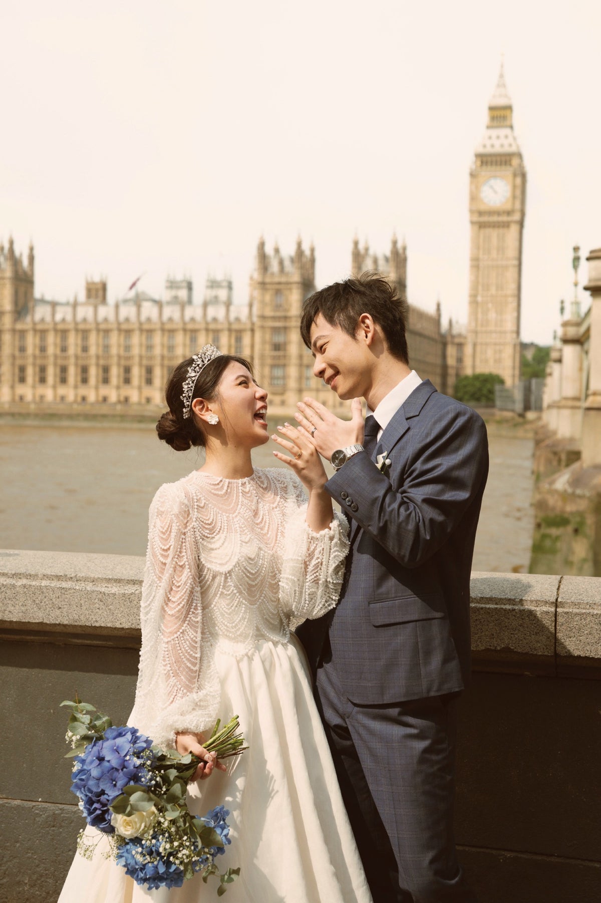 A bride in a white lace wedding gown holding a blue and white bouquet stands beside a groom in a navy suit in front of Big Ben and the Houses of Parliament on the Thames.