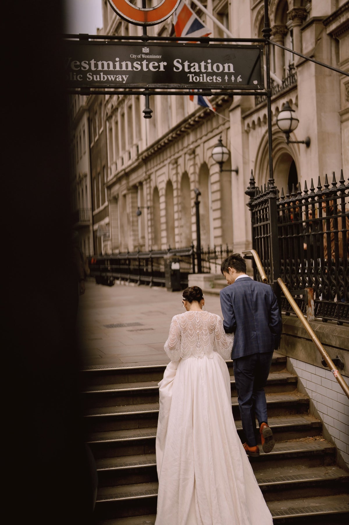 A bride in a white lace wedding gown and a groom in a navy suit walk down the stone steps of Westminster Station, viewed from behind through a dark foreground frame.