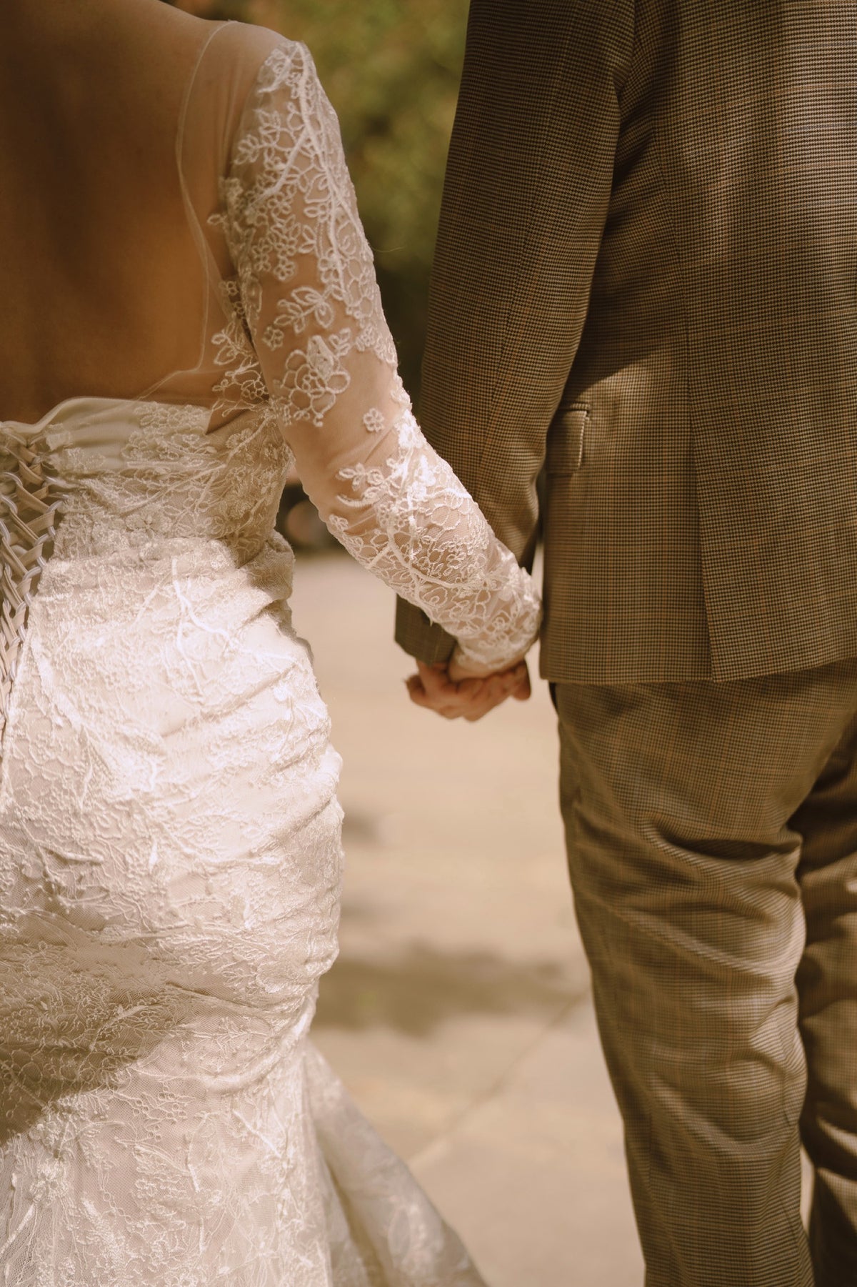 A close-up, over-the-shoulder view of a bride in a white lace wedding gown holding hands with a man wearing a brown plaid suit jacket and trousers, walking outdoors.