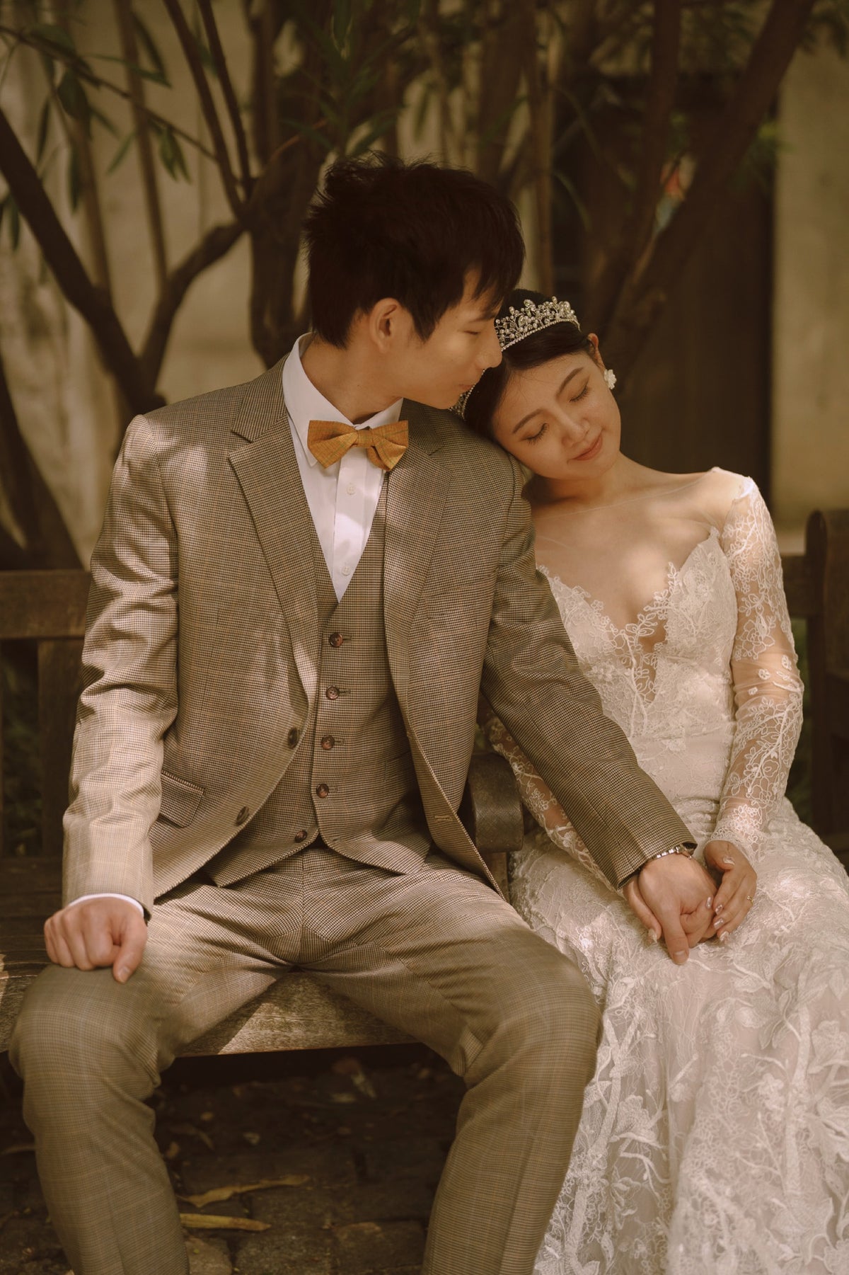 A couple in formal wedding attire sitting together on a bench outdoors. The groom wears a light taupe-beige three-piece suit with a yellow bow tie, and the bride wears a white lace wedding gown with a tiara.