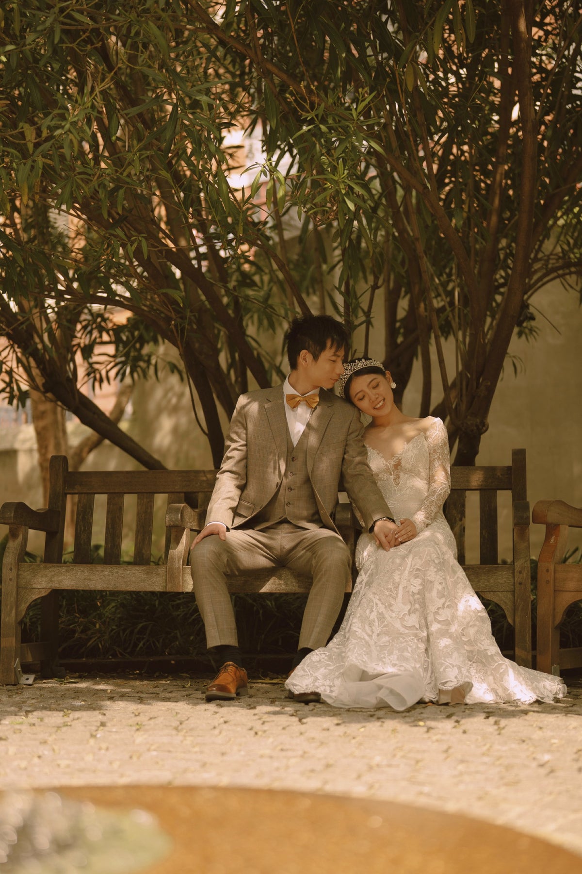 A bride in a white lace wedding gown and a groom in a beige suit sit together on a wooden bench under leafy trees, with warm sunlight filtering through the foliage.