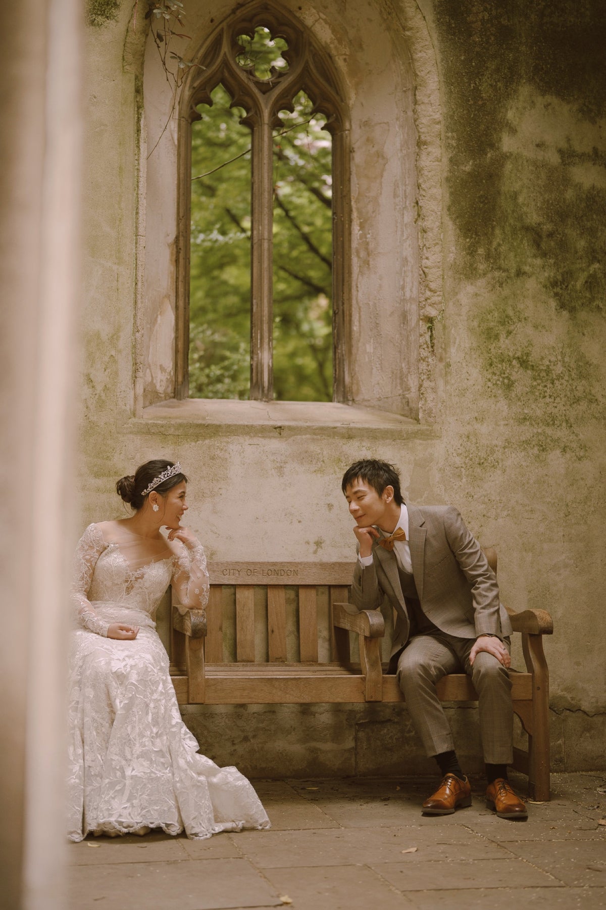 A bride in a white lace wedding gown sits on a bench next to a man in a gray suit, in front of a stone wall with a large arched window.