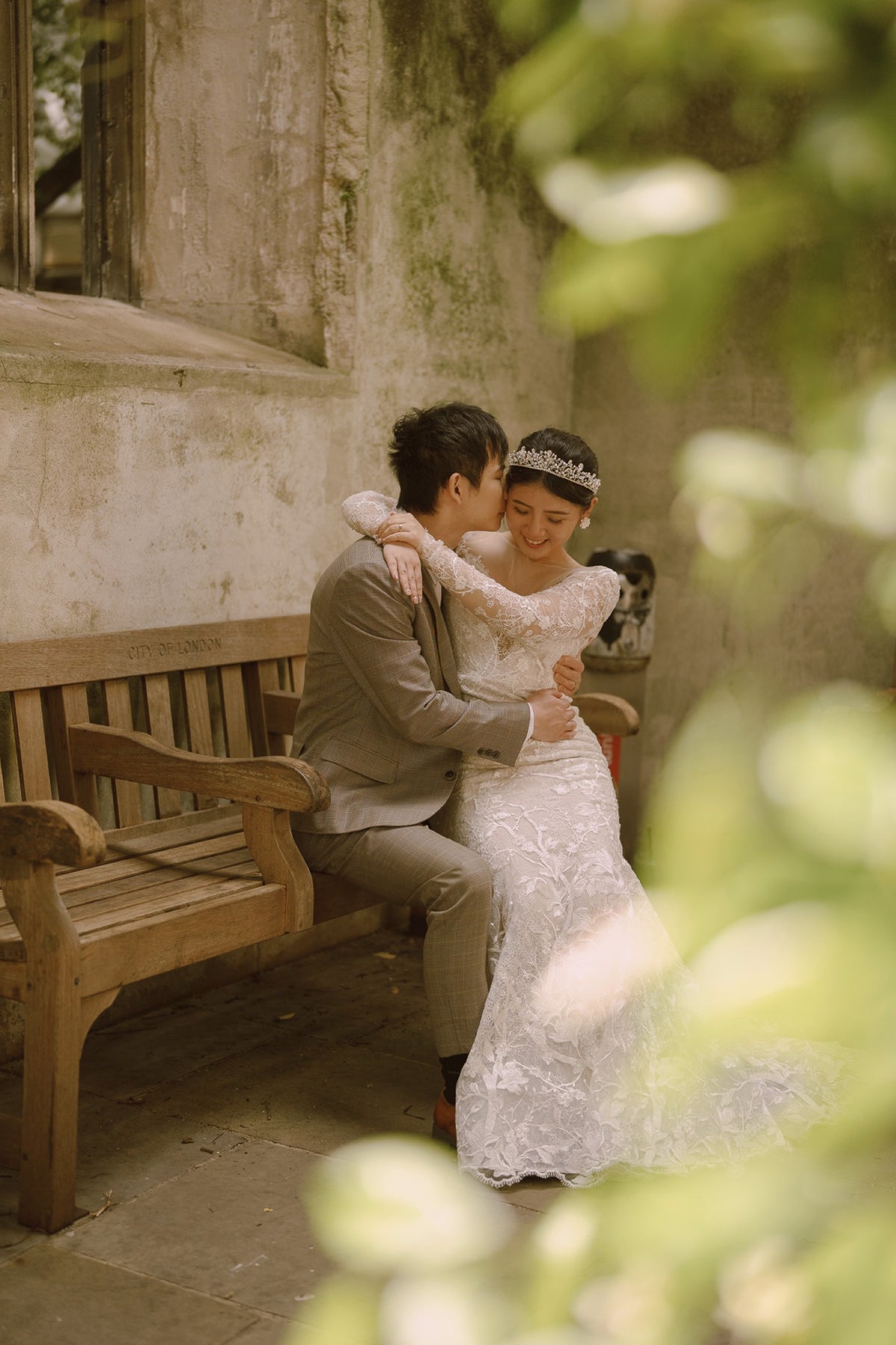 A bride in a white lace wedding gown and tiara embraces a groom in a taupe suit sitting on a wooden bench, framed by soft green foliage in the foreground.