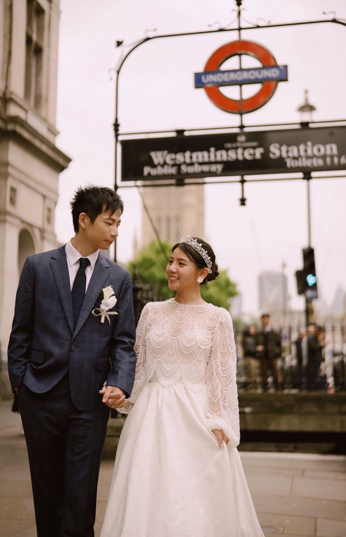 A bride in a white lace wedding gown and a groom in a navy blue suit holding hands, standing in front of a Westminster Station Underground sign on a city street.