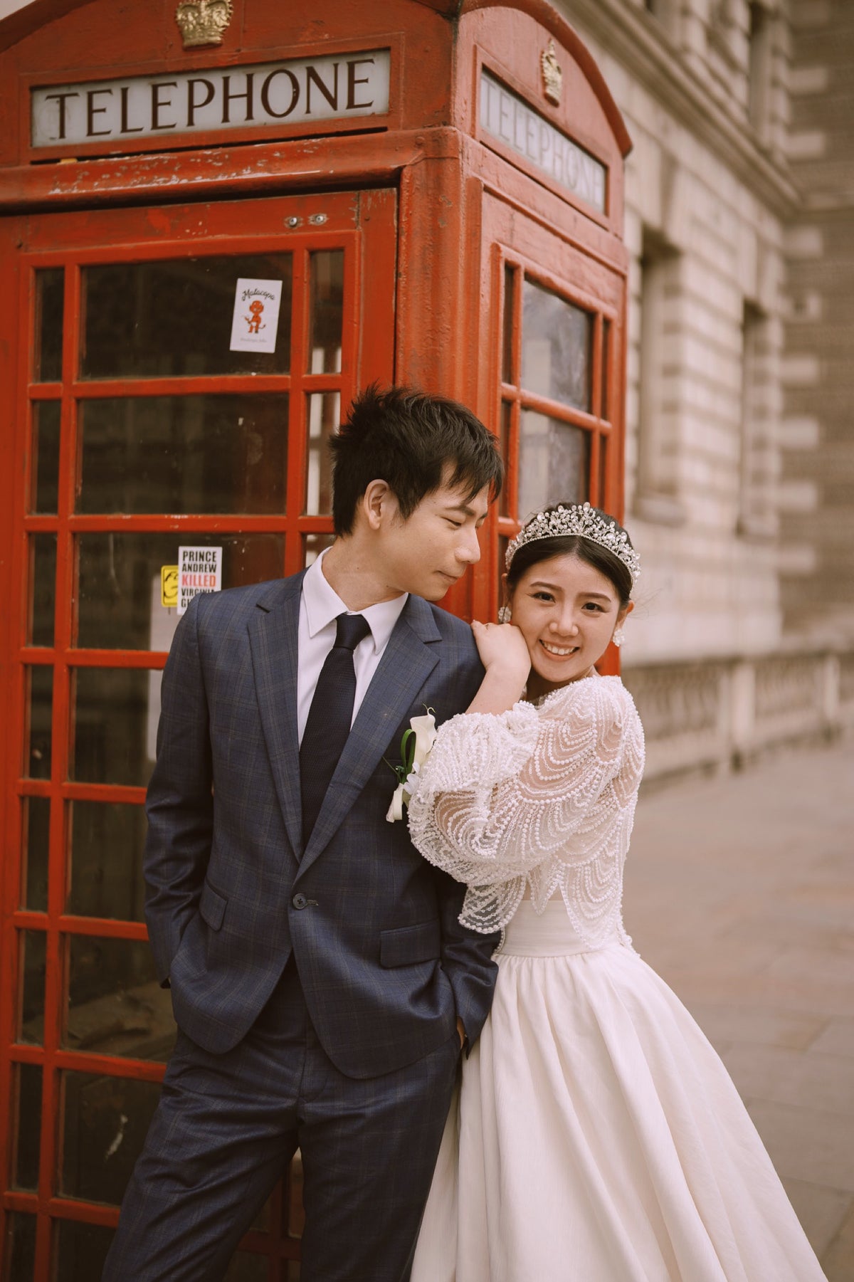 A bride in a white lace wedding gown and tiara stands beside a groom in a navy suit and tie, posing in front of a classic red telephone booth on a city street.