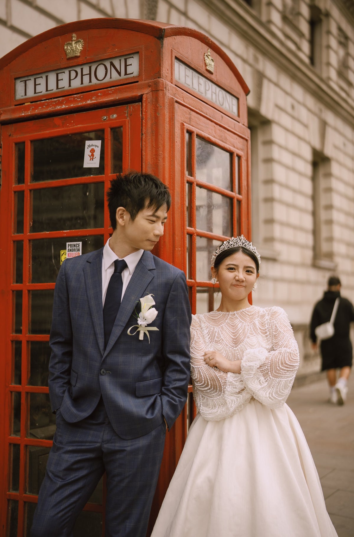 A couple posing outdoors in front of a classic red London telephone booth. The man wears a navy blue suit with a white rose boutonniere, and the woman wears a white lace wedding gown and tiara.