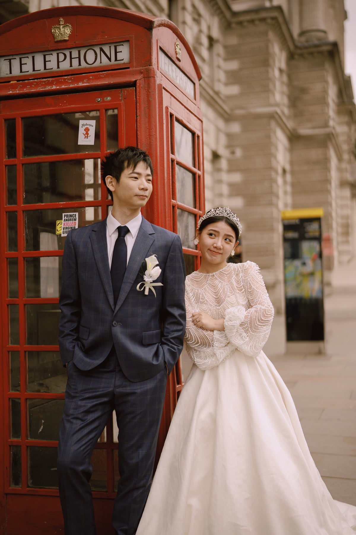 A bride in a white lace wedding gown and tiara stands beside a groom in a navy blue suit and tie, posing in front of a classic red London telephone booth and stone building.
