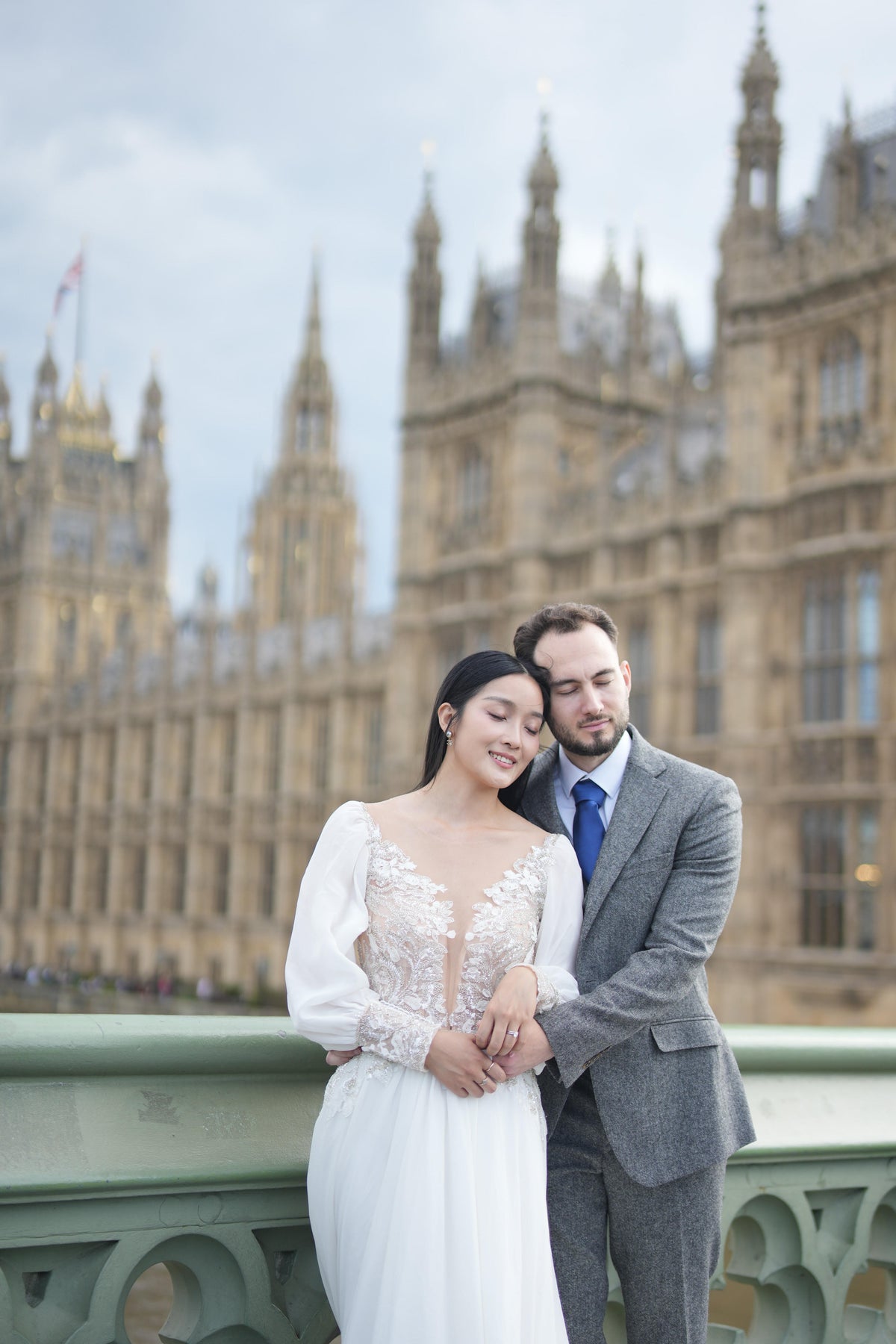 A bride in a white lace wedding gown and a groom in a gray suit and blue tie pose together in front of the Palace of Westminster and Big Ben in London.