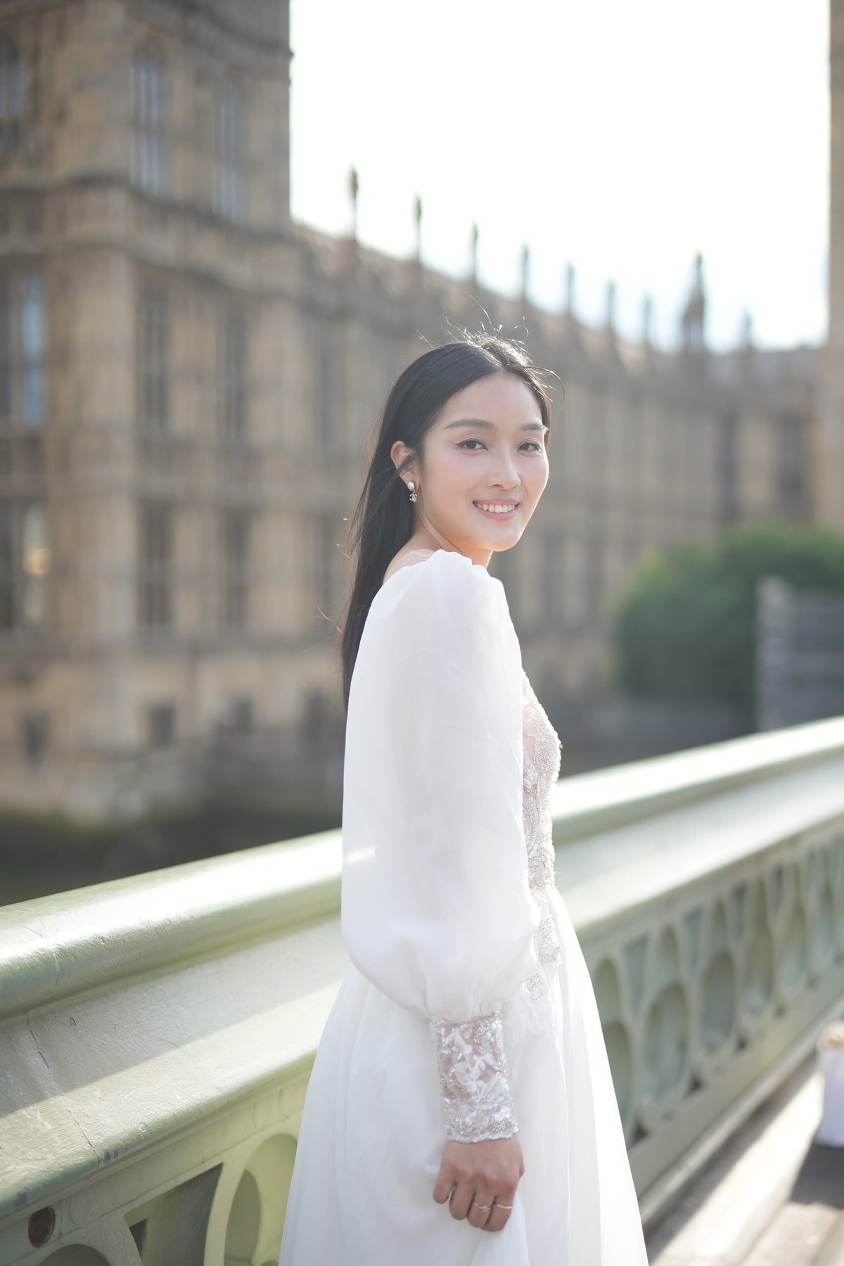 A woman wearing a white long-sleeve lace dress stands on a bridge with a large historic stone building in the background.