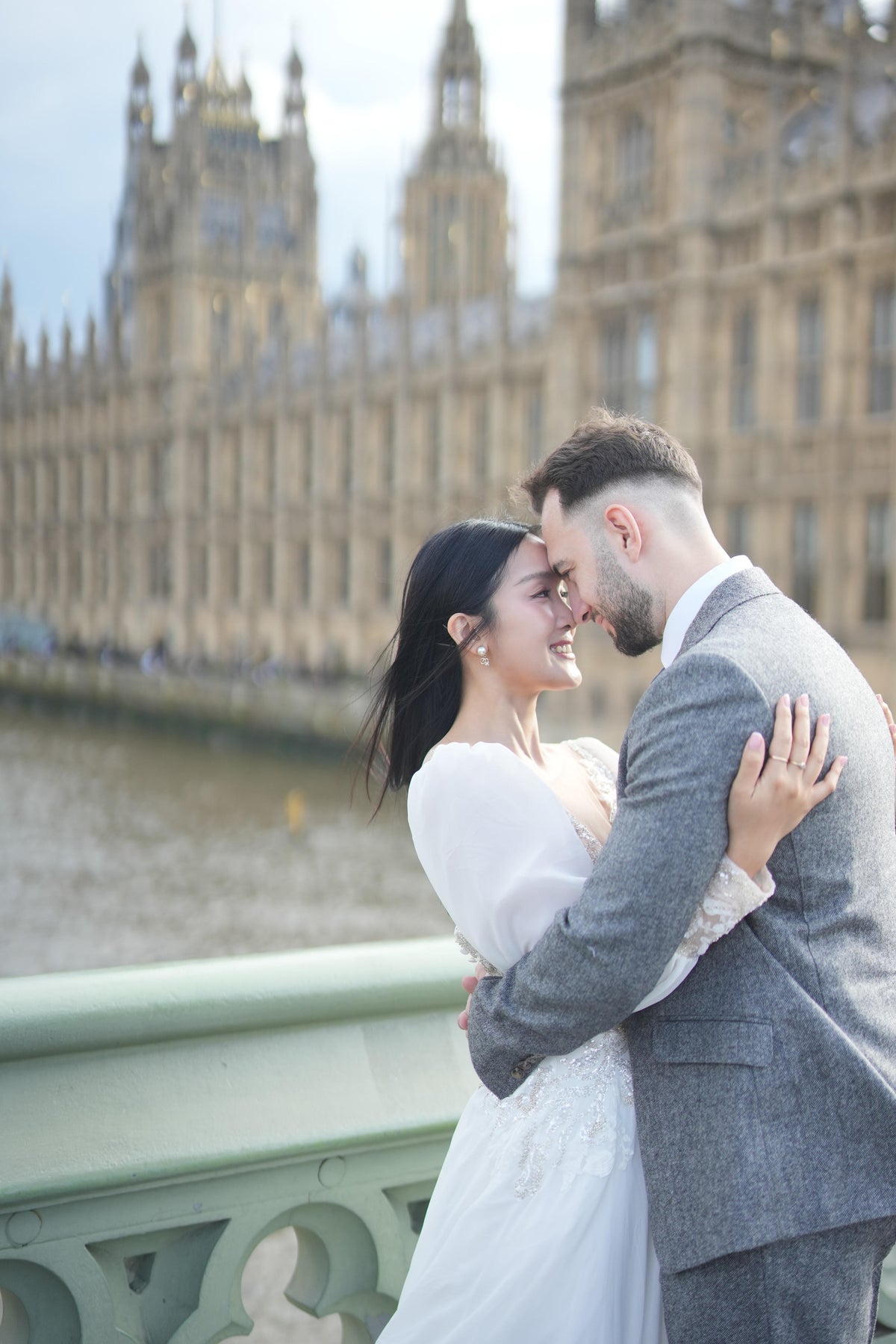 A bride in a white wedding dress and a groom in a gray suit embrace on Westminster Bridge with the Palace of Westminster and River Thames in London in the background.