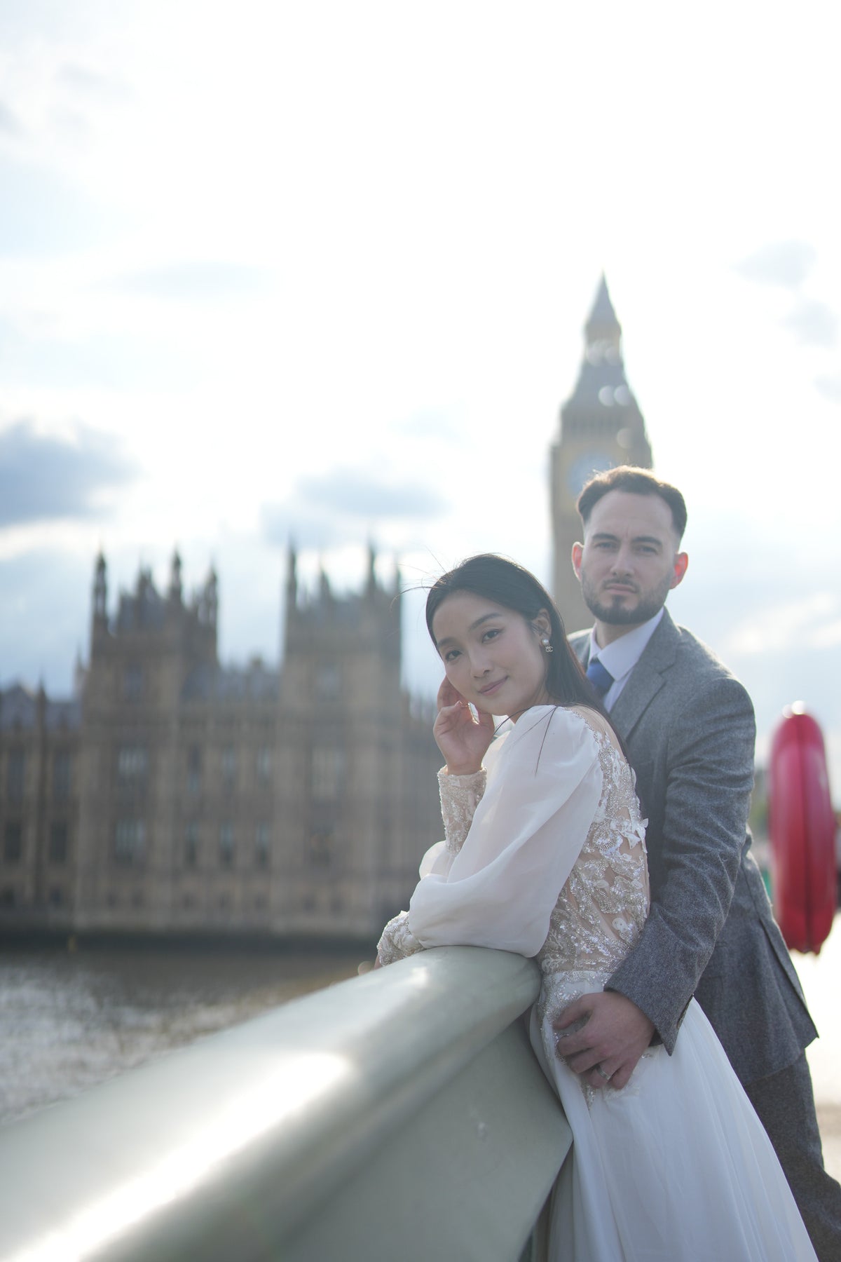 A bride in a white lace wedding gown and a groom in a gray suit pose together on a bridge with London's Big Ben and Houses of Parliament in the background.