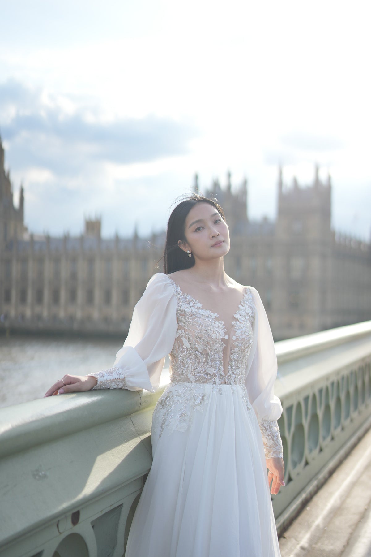 A woman wearing a white lace wedding gown stands on a bridge with a blurred London backdrop featuring the Houses of Parliament and river water.