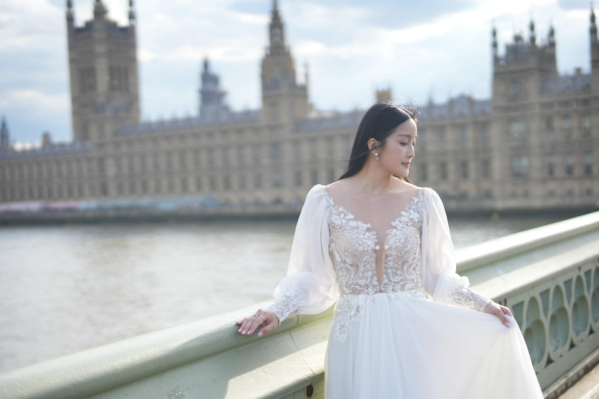 A woman wearing a white lace wedding gown stands on a bridge with the London Palace of Westminster and River Thames in the background.