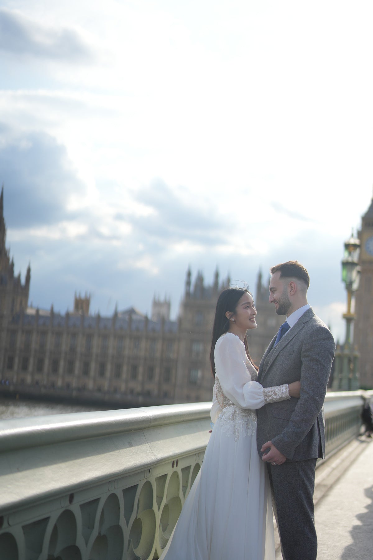 A bride in a white wedding gown and a groom in a gray suit standing together on Westminster Bridge with the Palace of Westminster and London Eye in the background.