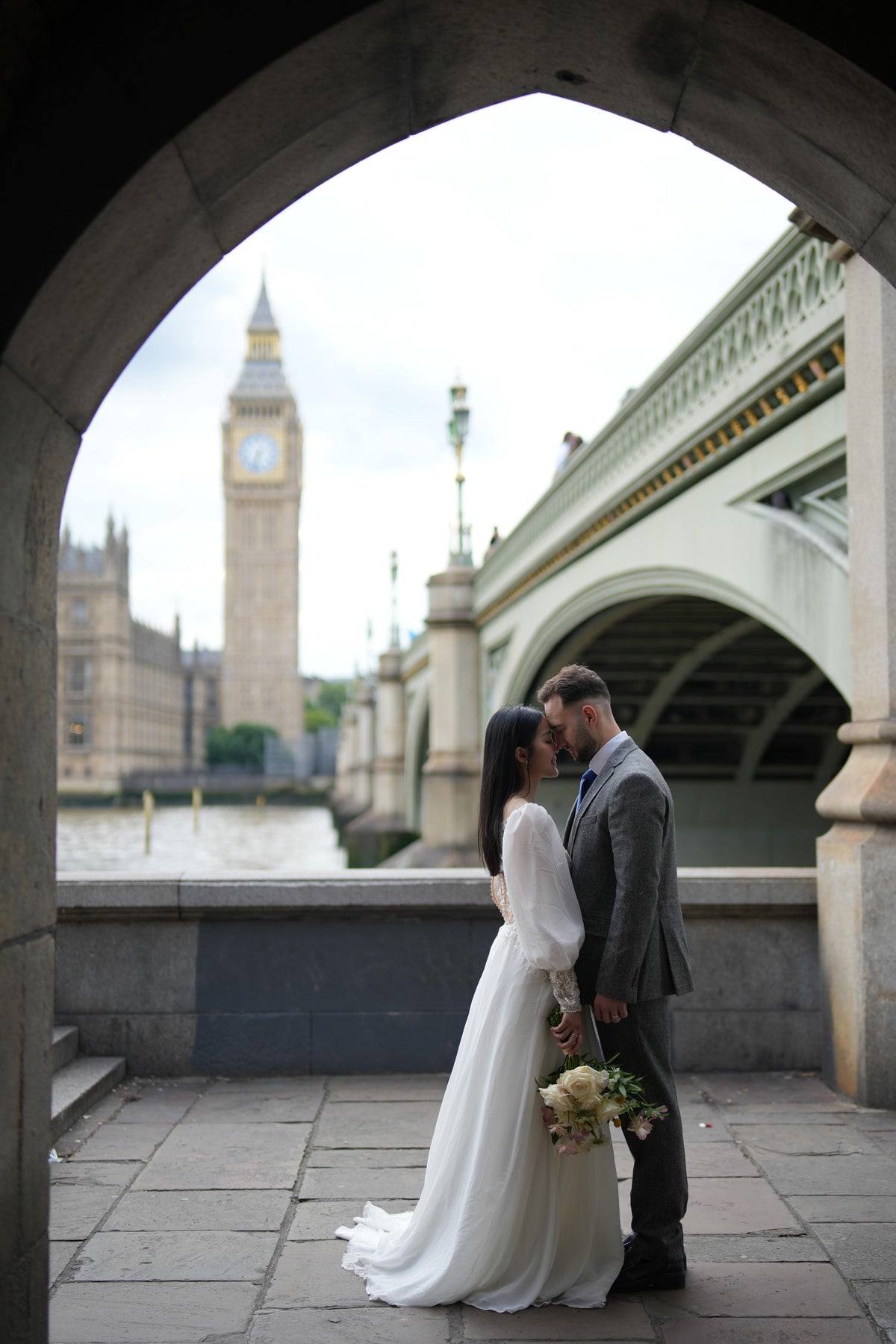 A bride in a white wedding gown and a groom in a gray suit kiss under a stone archway, with Big Ben and Westminster Bridge in London in the background.