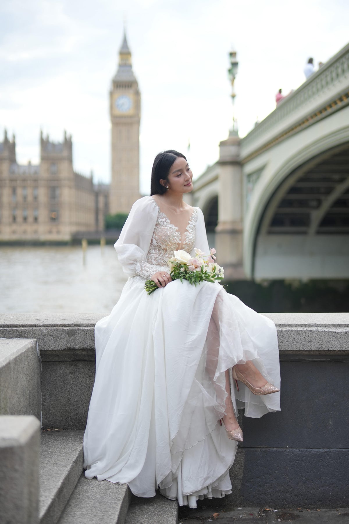 A woman in a white lace and tulle wedding gown sits on stone steps outdoors, holding a bouquet, with a river, bridge, and a clock tower in the background.