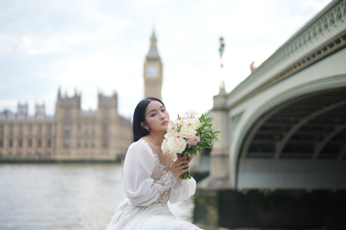 A woman in a white lace dress holding a bouquet of white and pale pink flowers, posed outdoors near water with a blurred London backdrop featuring Big Ben and Westminster Bridge.