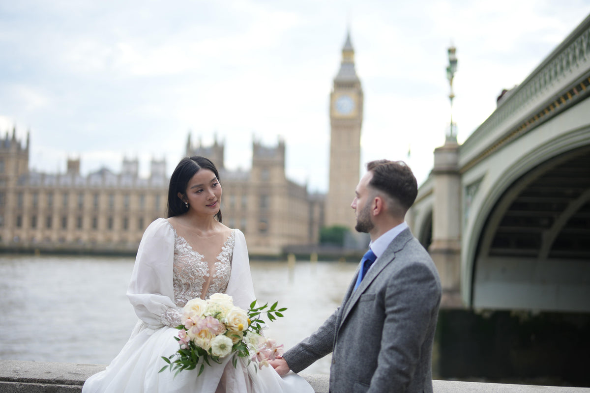 A bride in a white lace wedding gown holding a bouquet of white and pink flowers stands beside a groom in a gray suit on a riverside promenade, with Big Ben and Westminster Bridge in the background.