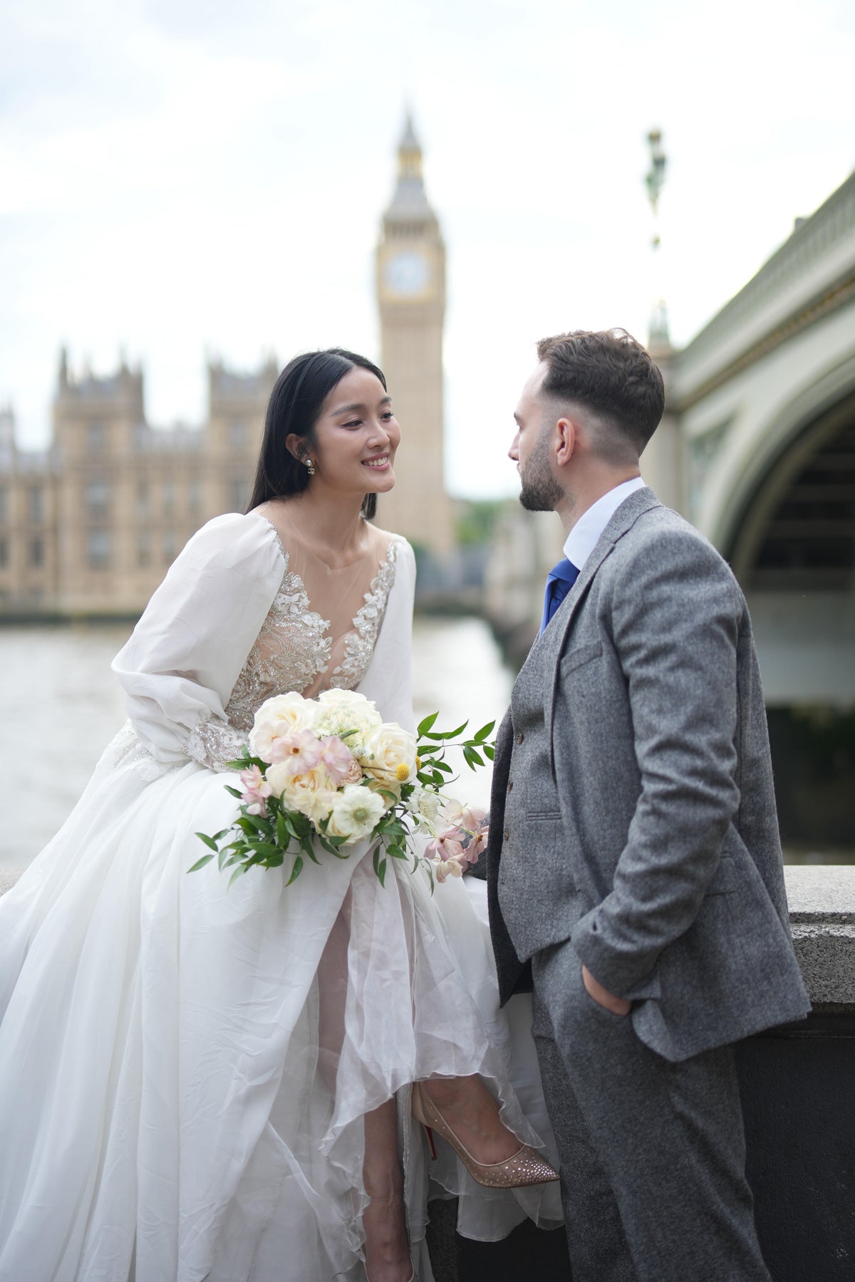 A bride in a white lace wedding gown holding a bouquet of white and pink flowers stands beside a groom in a gray suit, with Big Ben and a bridge blurred in the background.