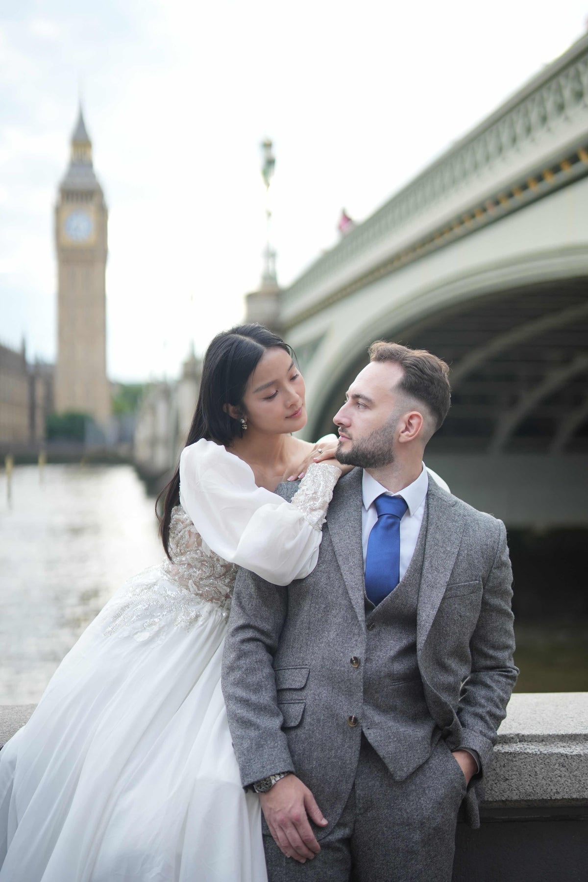 A bride in a white lace wedding gown and a groom in a gray three-piece suit with a blue tie pose together on a bridge, with Big Ben and the river visible in the background.