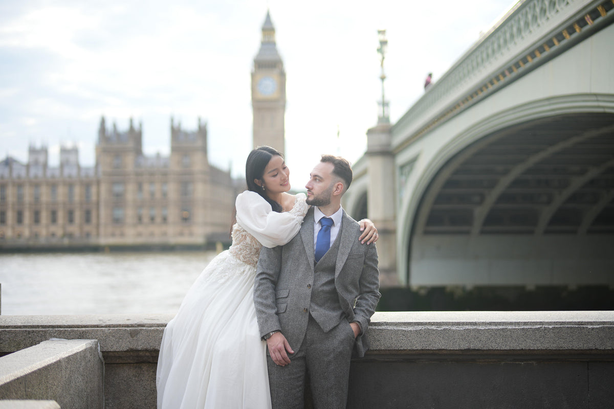 A bride in a white wedding gown and a groom in a gray suit pose together on a bridge in London, with Big Ben and Westminster Bridge in the background.