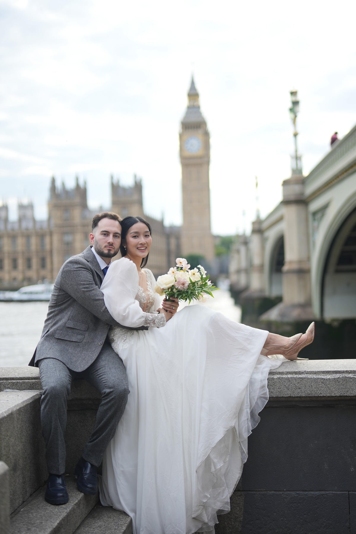 A bride in a white wedding gown and a groom in a gray suit sit together on stone steps in London, with Big Ben and the Houses of Parliament in the background.