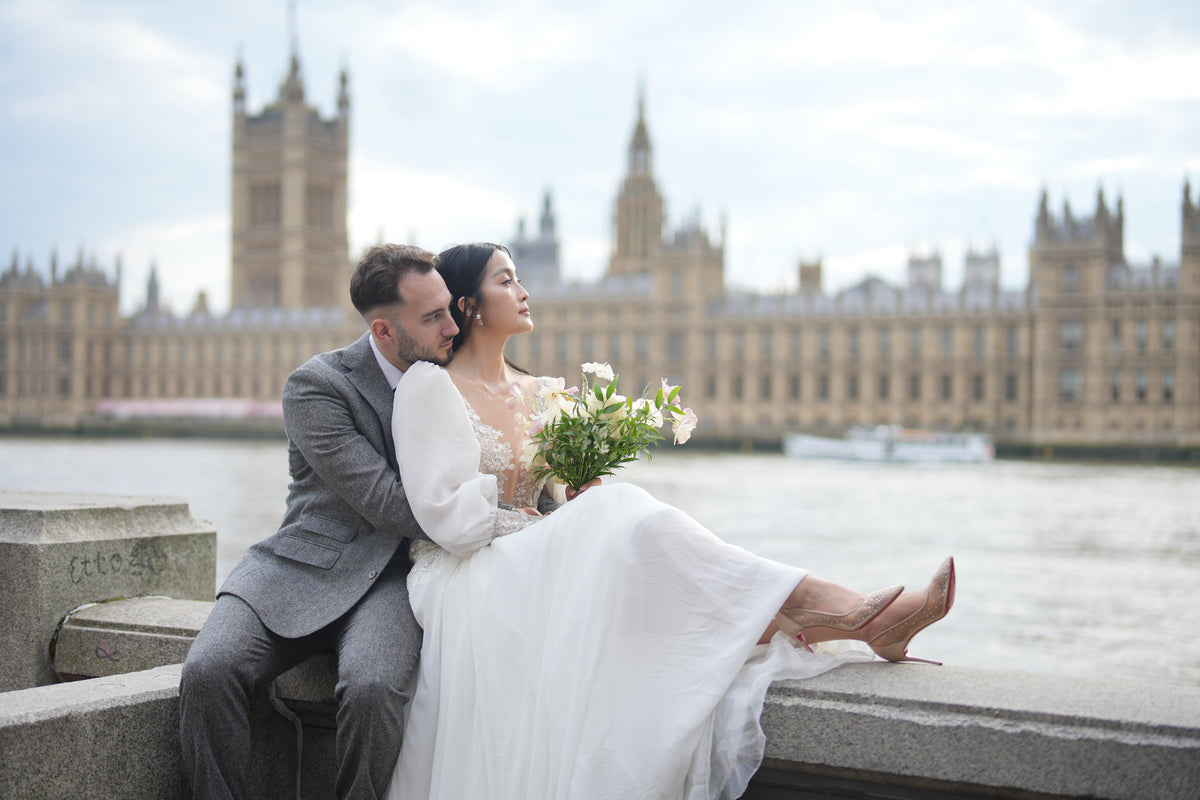 A bride in a white wedding gown and a groom in a gray suit sit together on a stone embankment in London, with the Houses of Parliament and River Thames in the background.