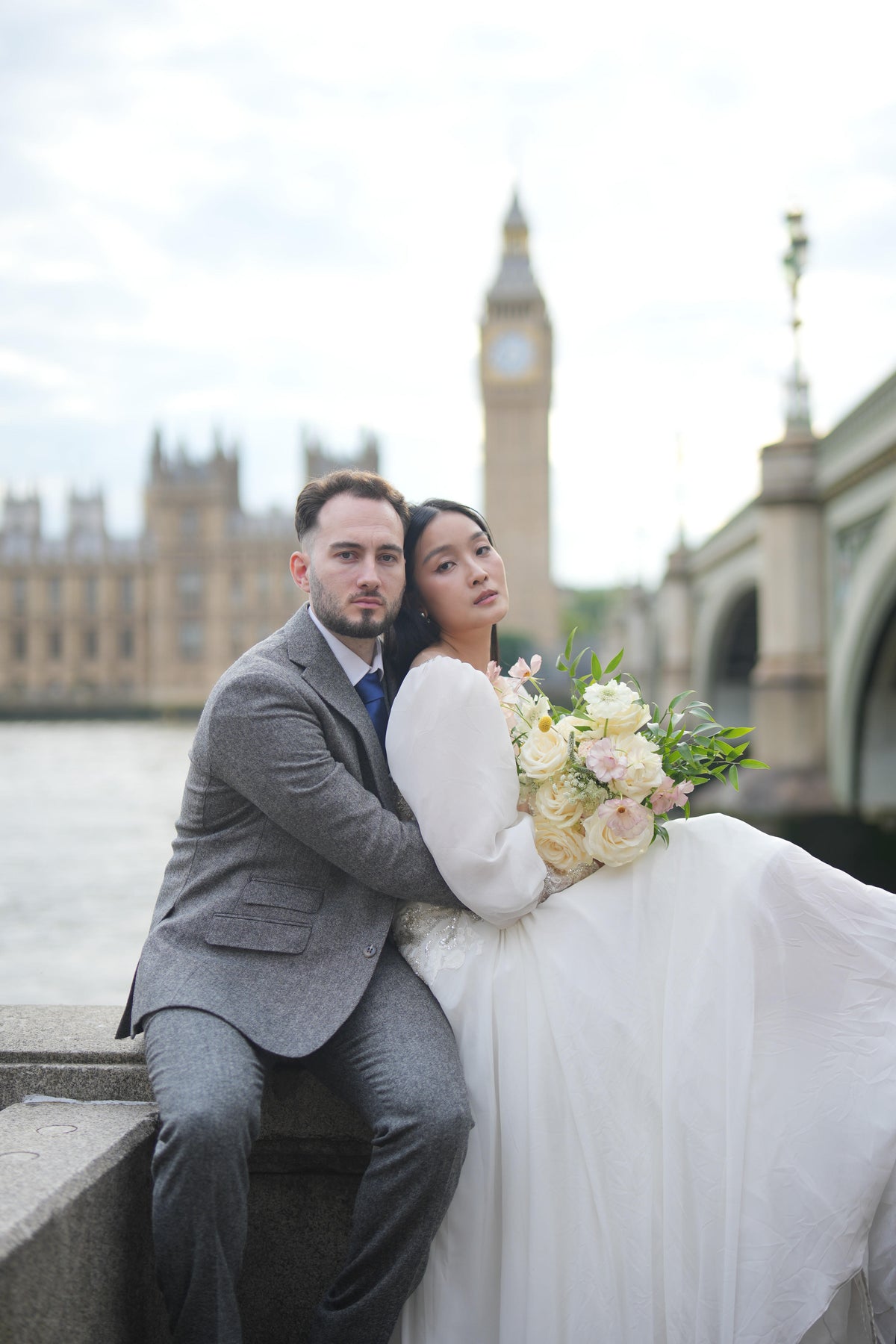 A bride in a white wedding gown and a groom in a gray suit sit together on a stone ledge in London, with Big Ben and the Houses of Parliament blurred in the background.
