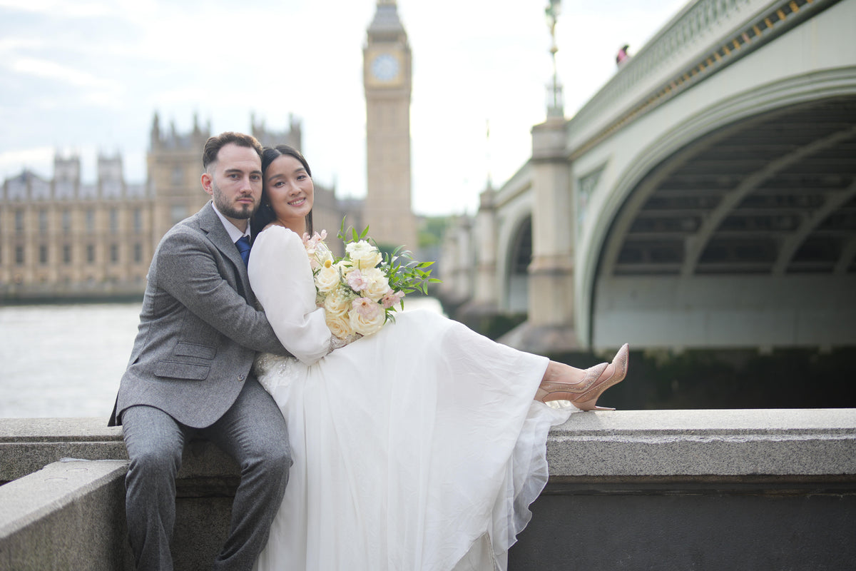 A bride in a white wedding gown and a groom in a gray suit sit together on a stone bridge in London, with Big Ben and Westminster Bridge in the background.