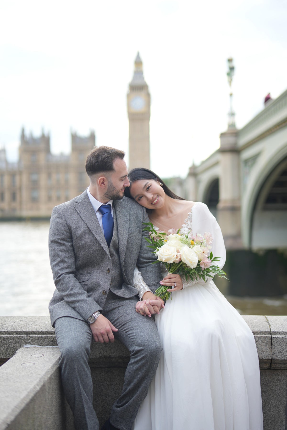 A bride in a white wedding gown and a man in a gray suit sit together on a stone ledge with Big Ben and Westminster Bridge in the background.