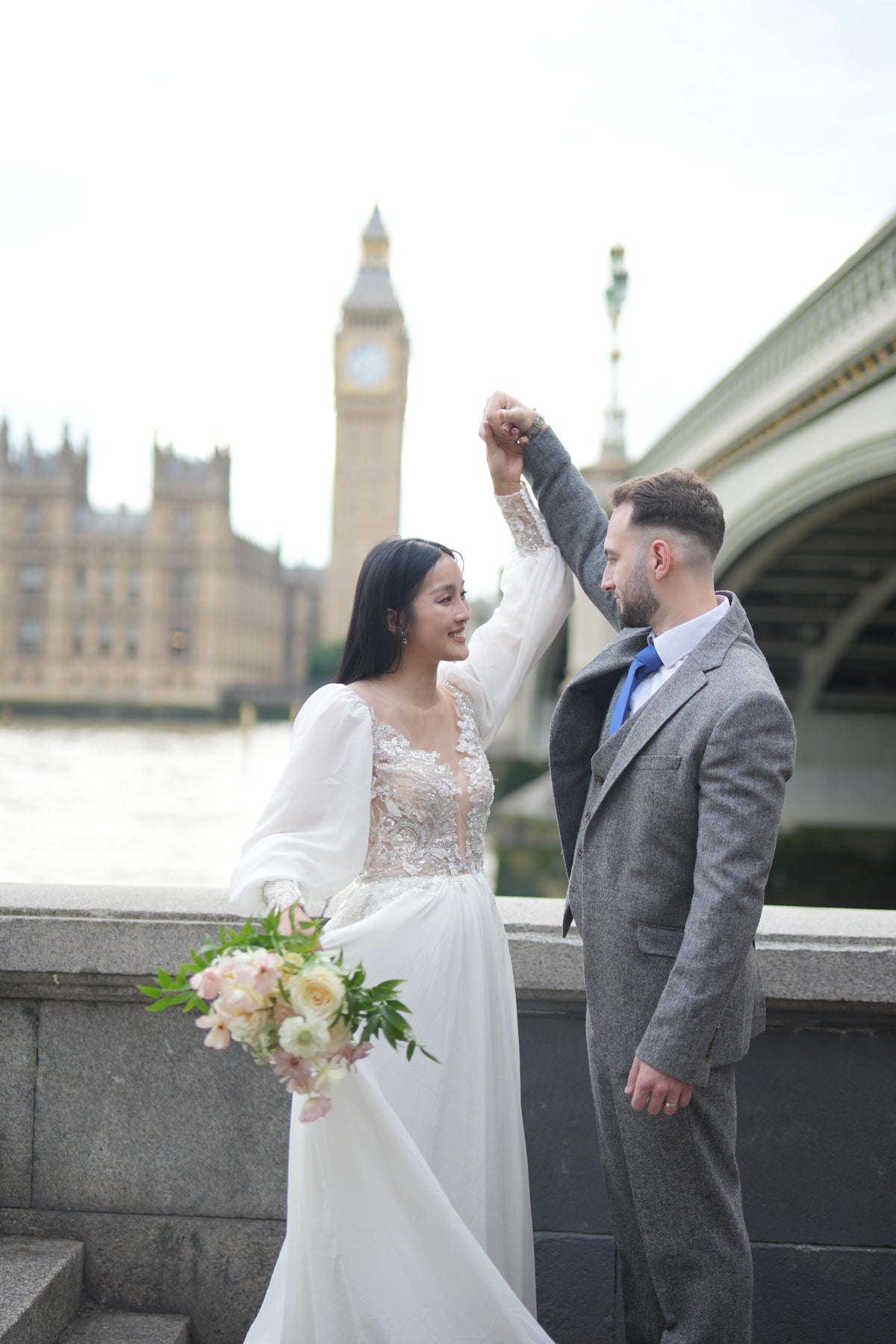 A bride in a white lace wedding gown holding a bouquet of pale pink flowers and dancing with a groom in a gray suit on a bridge, with Big Ben and Westminster Bridge in London in the background.