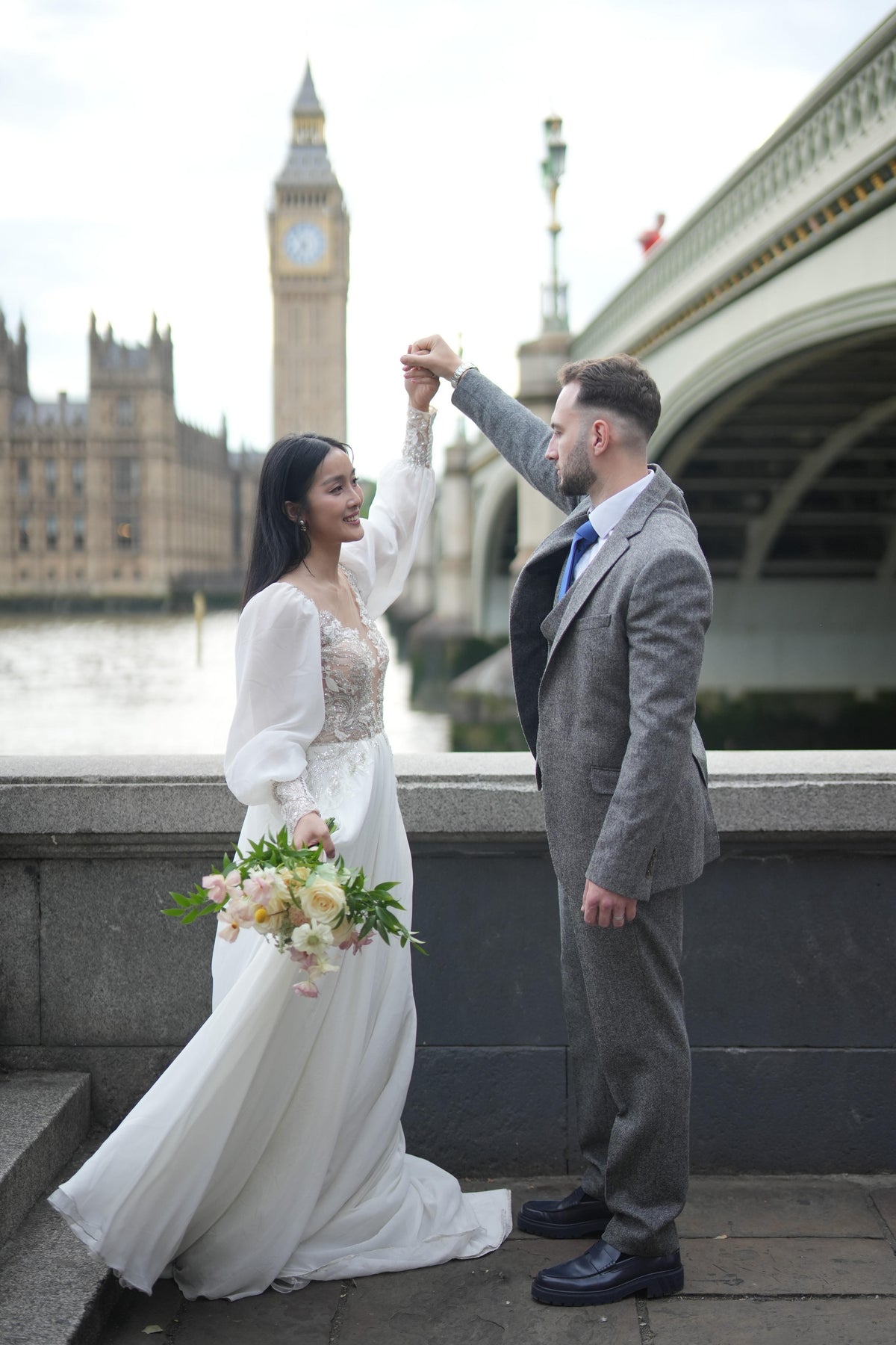 A bride in a white long-sleeve wedding gown holding a bouquet of pink and white flowers stands on a bridge next to a man in a gray suit, with Big Ben and Westminster Bridge in London in the background.