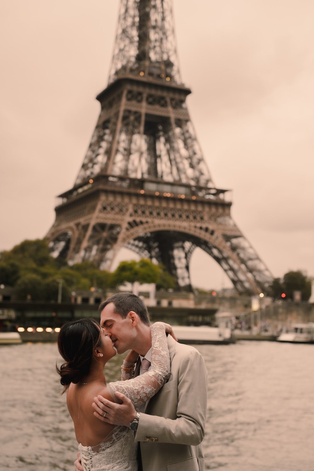 A couple embracing and kissing in front of the Eiffel Tower in Paris, with a river and boats in the background. The woman wears a white lace-trimmed dress, and the man wears a light beige suit.