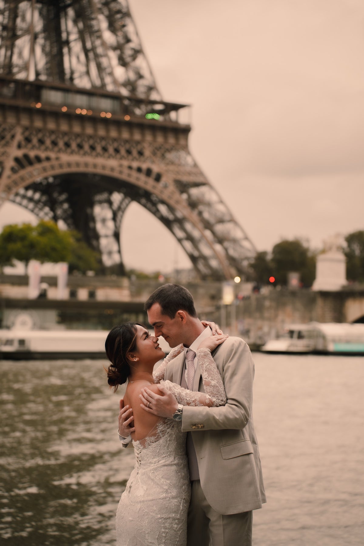A bride in a white lace wedding gown and a groom in a light gray suit embrace and kiss in front of the Eiffel Tower, with the Seine River and boats in the background.