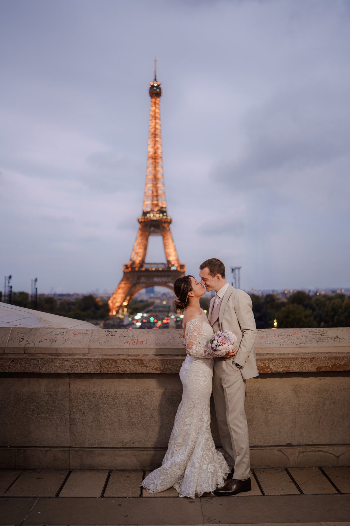 A bride in a white lace wedding gown and a groom in a light beige suit kiss in front of the illuminated Eiffel Tower at dusk, with a stone wall and city skyline in the background.