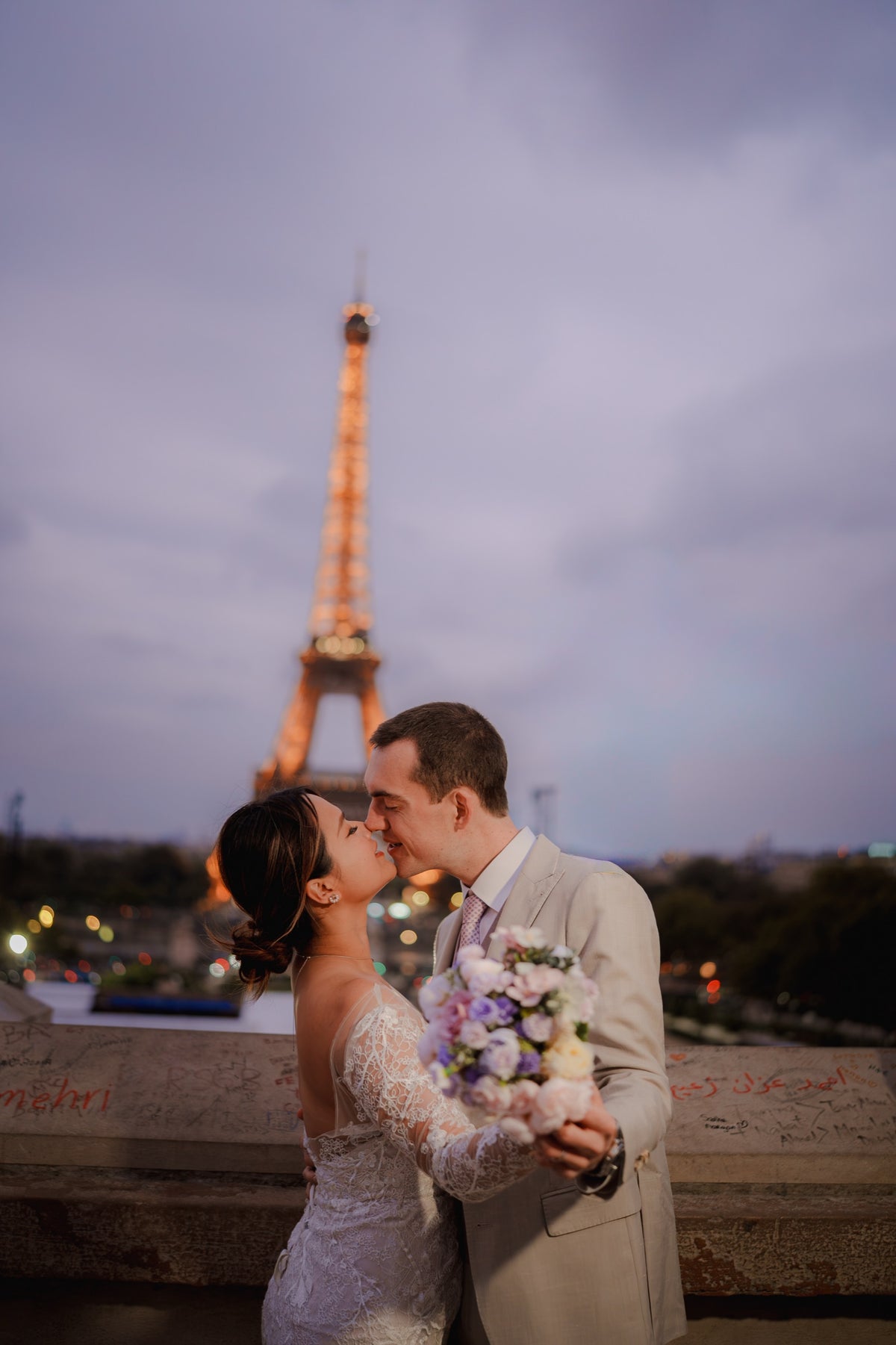 A bride in a white lace wedding gown and a groom in a light beige suit kiss in front of the illuminated Eiffel Tower at dusk, with the bride holding a pastel bouquet.