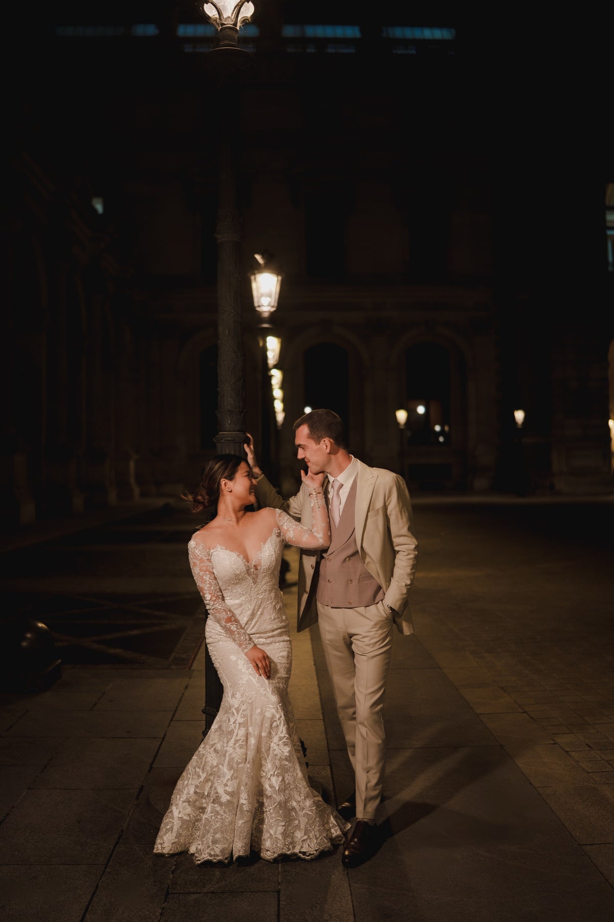 A bride in a strapless lace mermaid gown and a groom in a light beige suit pose together on a cobblestone street at night under warm streetlights.