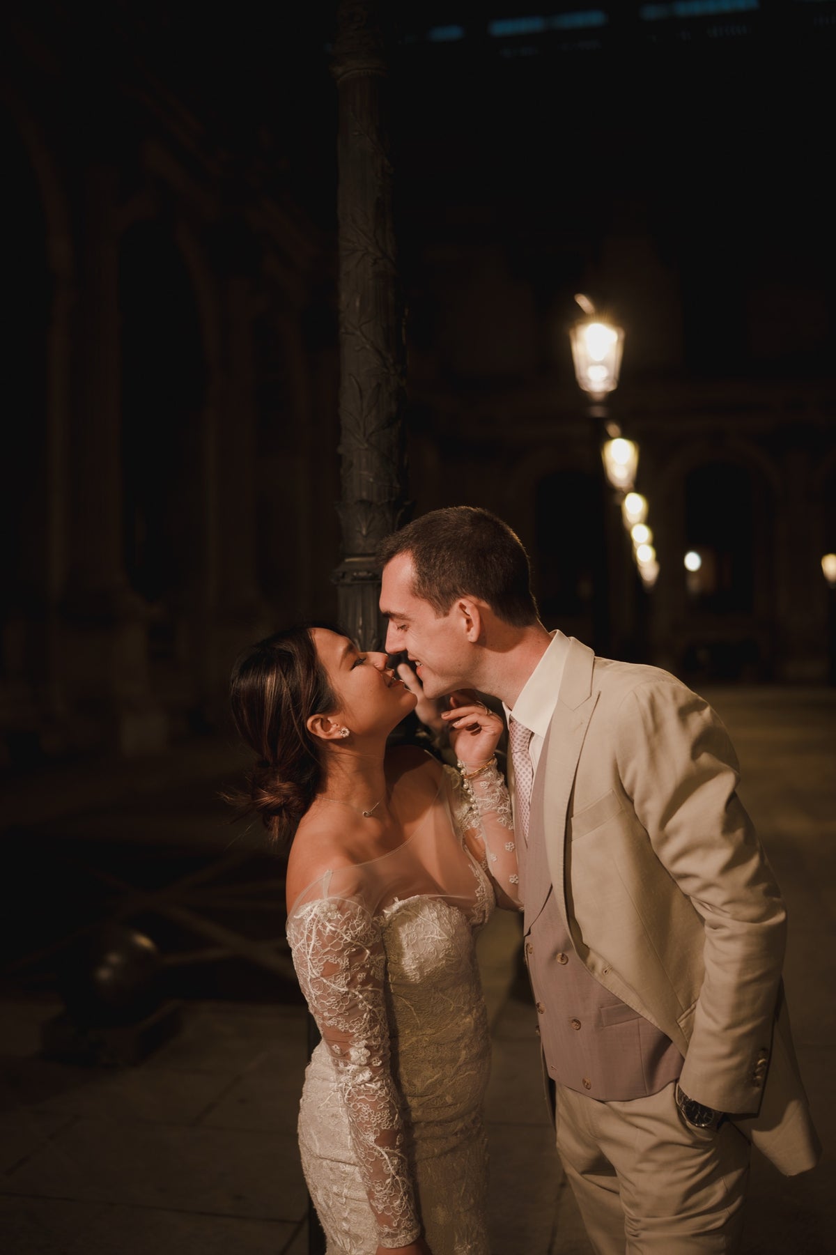 A bride in a strapless lace wedding gown and a groom in a light beige suit embrace and kiss in a dimly lit outdoor night setting with warm streetlights in the background.