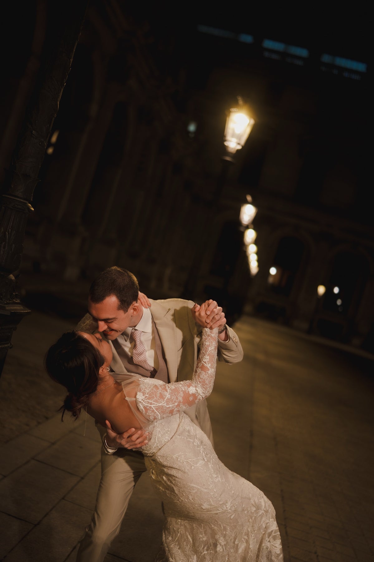 A bride in a long-sleeve lace wedding gown and a groom in a beige suit embrace and dance on a dimly lit cobblestone street at night.