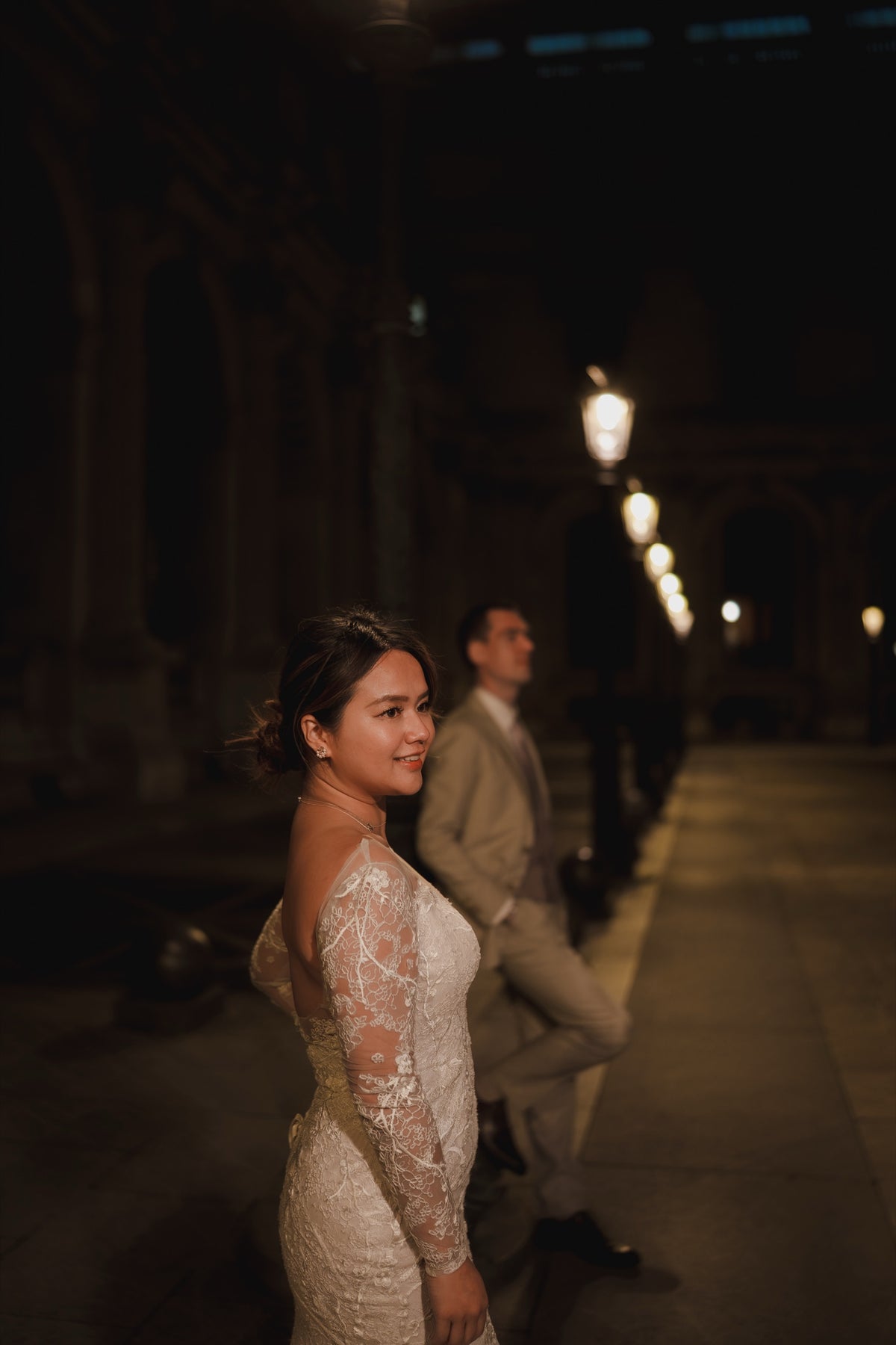 A woman in a fitted lace wedding gown stands in a dimly lit nighttime corridor, looking back over her shoulder. A man in a light beige suit is blurred in the background near glowing street lamps.