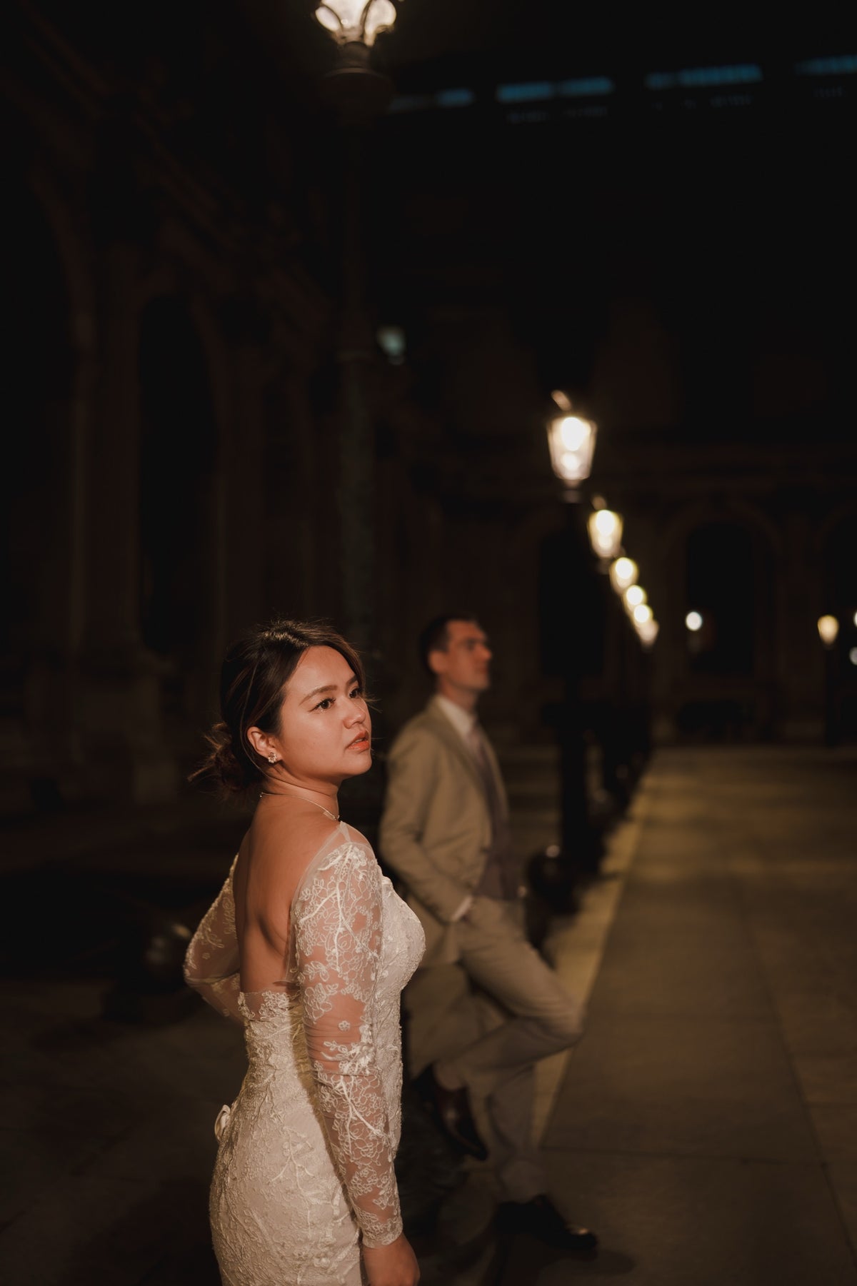 A bride in a white lace wedding gown stands in a dimly lit historic corridor at night, looking back over her shoulder. A groom in a light beige suit is visible in the background under warm streetlamp light.
